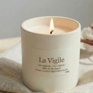 Bougie parfumée La Vigile de Bain de Cire, notes de cuir patiné et miel de montagne, en céramique blanche avec flamme allumée dans une ambiance douce.