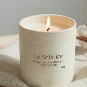 Bougie allumée Le Solstice par Bain de Cire, senteur fleur d'oranger et santal, en pot de céramique blanche posé sur un tissu en lin naturel.